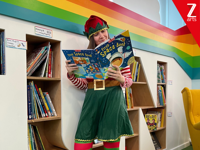 A storyteller is dressed up as an elf as they read a book in the library.