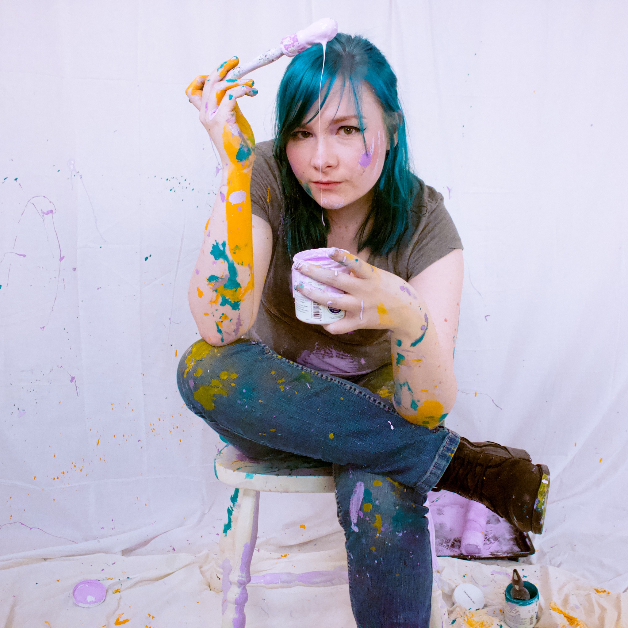 A musician sits on a stool holding a paint pot in one hand and a paintbrush on top of her head in the other.