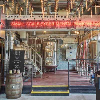 Inside in an industrial building with a red neon light that reads 'small scale experiemental beer machine'.
