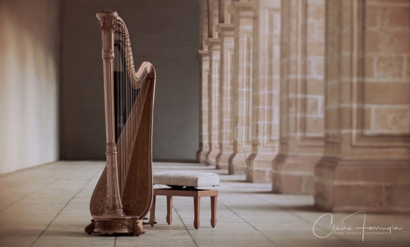 A wooden harp with a stool next to it in an ornate trevantine hall with tall pillars running down the right side of it.