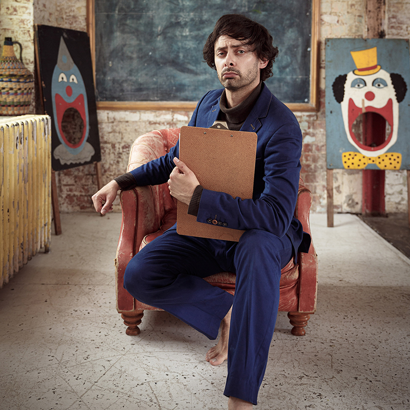 French comedian Marcel Lucont sits on a brown chair wearing a blue suit and holding a clipboard, with a serious expression on his face.There is a picture of a clown in the background.