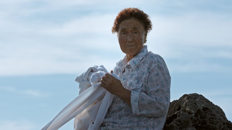 An elderly woman with short brown hair leans against a rock on Jeju Island, south of the Korean peninsula. Looking at the viewer with a neutral expression, she wears a long sleeved blouse with a floral pattern and is holding a long white cloth.