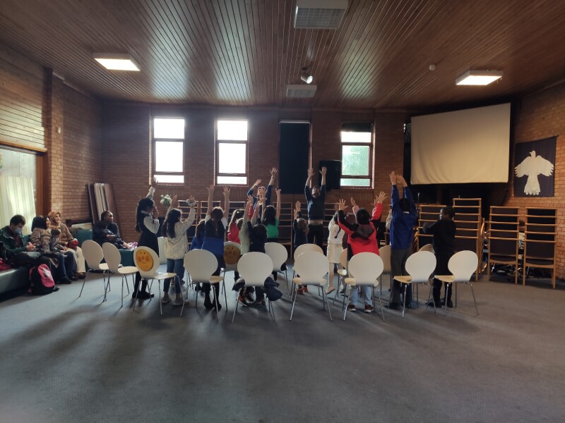 A community room with a group of people who are stood up and raising their arms up into the air. There is a circle of white chairs around them.