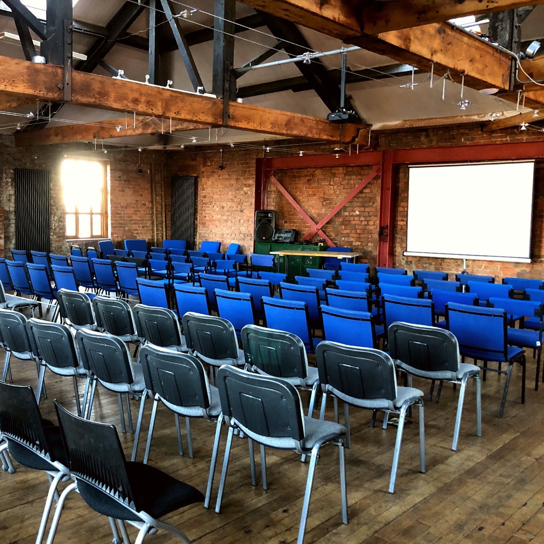 A room with a vaulted ceiling of dark wooden beams set out with rows of blue chairs.