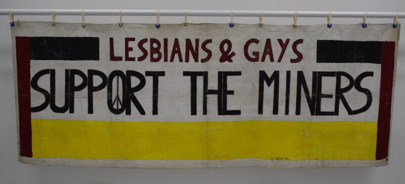 A white banner with black, red and yellow stripes. The text reads: Lesbians & Gays Support The Miners