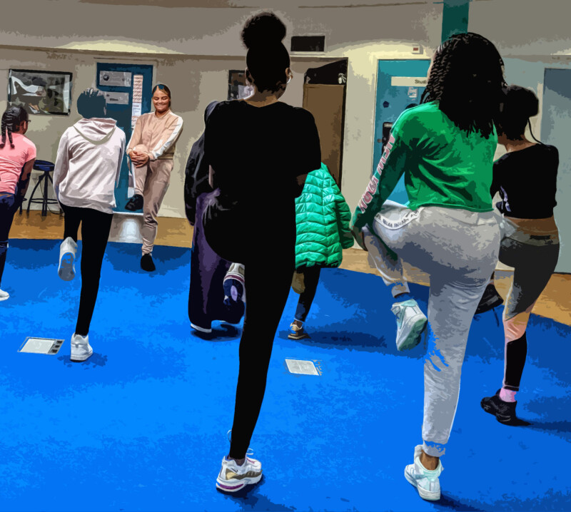 A group of young people doing a stretching exercise with their left leg lifted and bent at the knee, inside a dance studio with a blue floor.