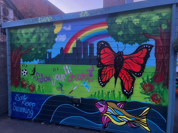An outside brick wall that has been decorated with an outdoor scene of grass, blue skies, trees and a butterfly.