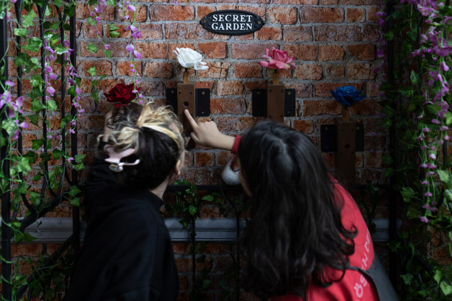 Two young people stand infront of a brick wall. One of the yung people points to the wall. There is a plaque on the wall that reads "Secret Garden".