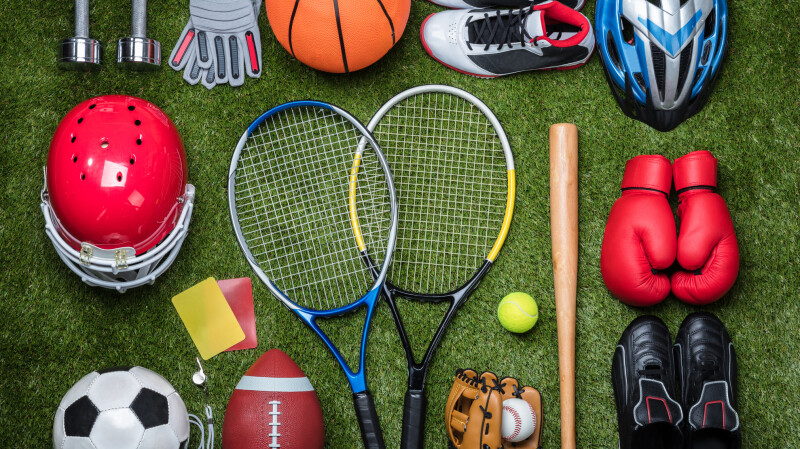 A selection of different sports equipment. There is a rugby ball, tennis rackets, a baseball, gloves and football boots.
