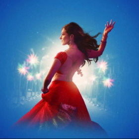 A beautiful female Indian dance stands wearing a red skirt and top with her right hand raised upwards. There are white sparkles around her on a blue background.