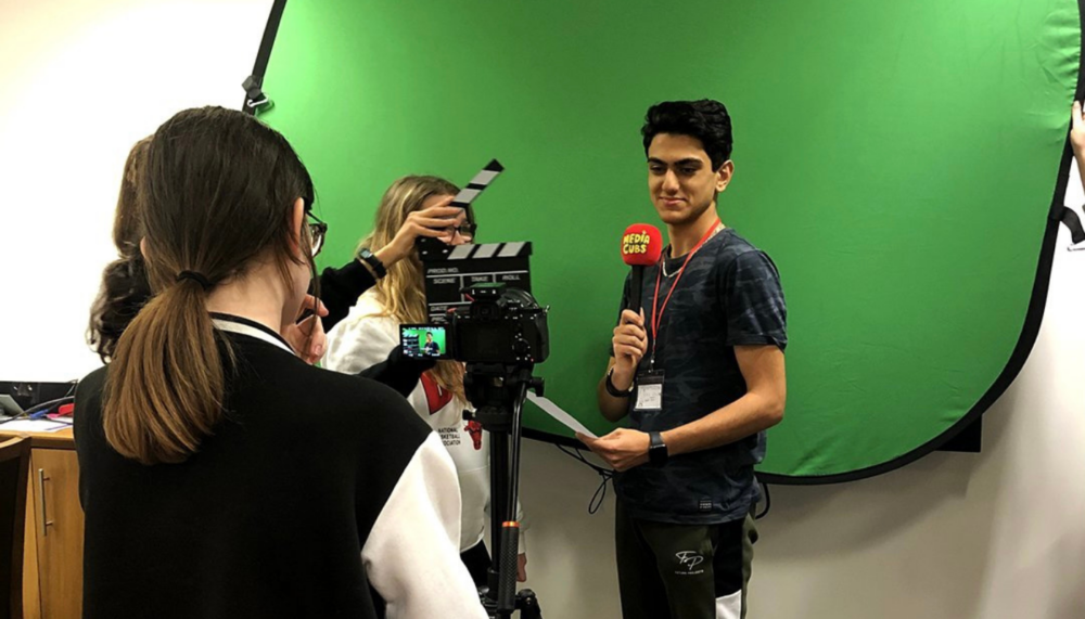 A young person stands in front of a green screen holding a microphone as another young person films them.