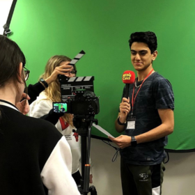 A young person stands in front of a green screen holding a microphone as another young person films them.
