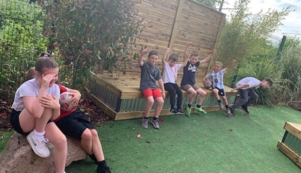 A group of children sat on a wooden bench outside with a grassy lawn in front of them.