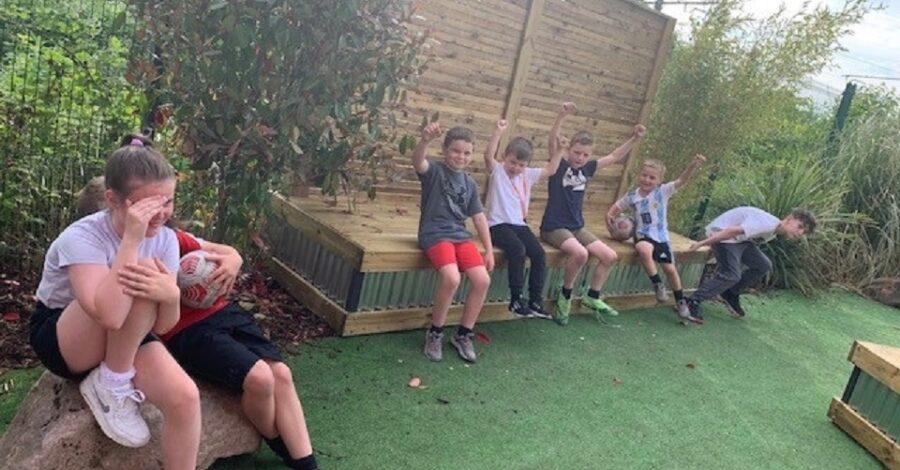A group of children sat on a wooden bench outside with a grassy lawn in front of them.