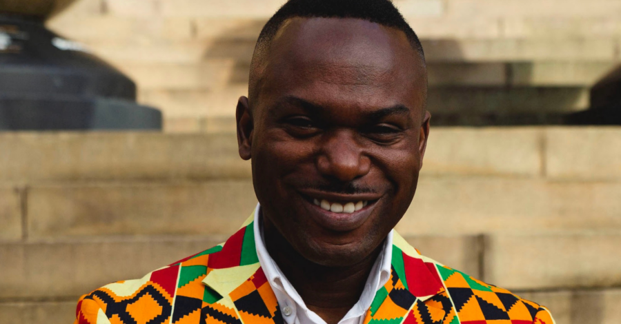 The head a shoulders of poet Jason Allen Paisant. He is wearing a brightly coloured jacket of orange, red, yellow and black over a white shirt.
