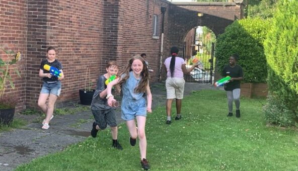 Three young people running and playing with water pistols outside. There is a tall brick wall to the side of them, a path and a grassy lawn.