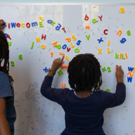 A small child stands facing a magnetic white board playing with magnetic brightly coloured letters.