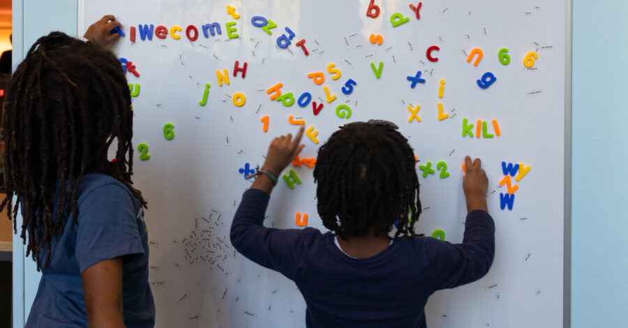 A small child stands facing a magnetic white board playing with magnetic brightly coloured letters.