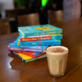 A pile of seven brightly coloured books and a coffee in a glass on a wooden table.