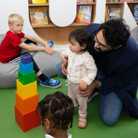 An adult kneels behind a toddler who is staring at a tower of brightly coloured blocks in front of them. Another small child sits on a grey beanbag next to them.