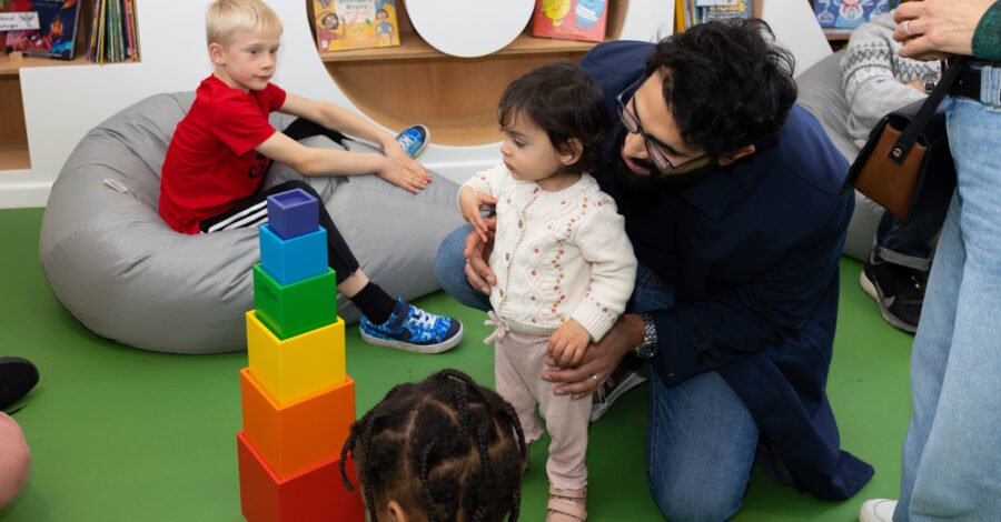 An adult kneels behind a toddler who is staring at a tower of brightly coloured blocks in front of them. Another small child sits on a grey beanbag next to them.