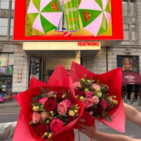 Two bouquets of red and pink flowers, wrapped in red paper held up in front of the Printworks entrance.