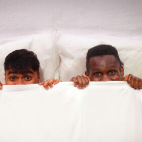 Two young children lie in a bed with a white duvet and white pillows. They grab the duvet and pull it up to underneath their noses.