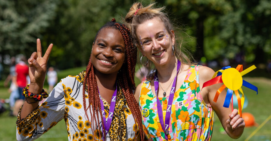 Two women stand close to oneanother wearing highly patterned, brightly coloured clothes smiling. They are standing on a grassy lawn in the day time with trees at the back of them.