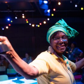 A lady with a yellow t-shirt and a green headscarf on holds her arms out as she dances.
