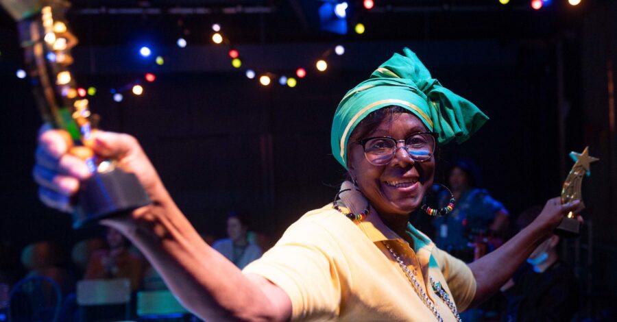 A lady with a yellow t-shirt and a green headscarf on holds her arms out as she dances.