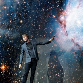 Professor Brian Cox stands on a stage with his left arm pointing to an image of space.