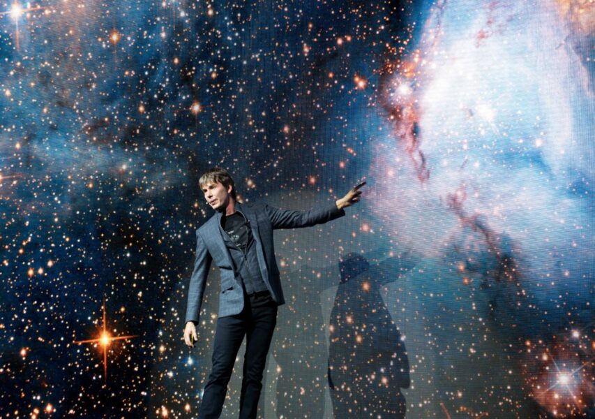 Professor Brian Cox stands on a stage with his left arm pointing to an image of space.