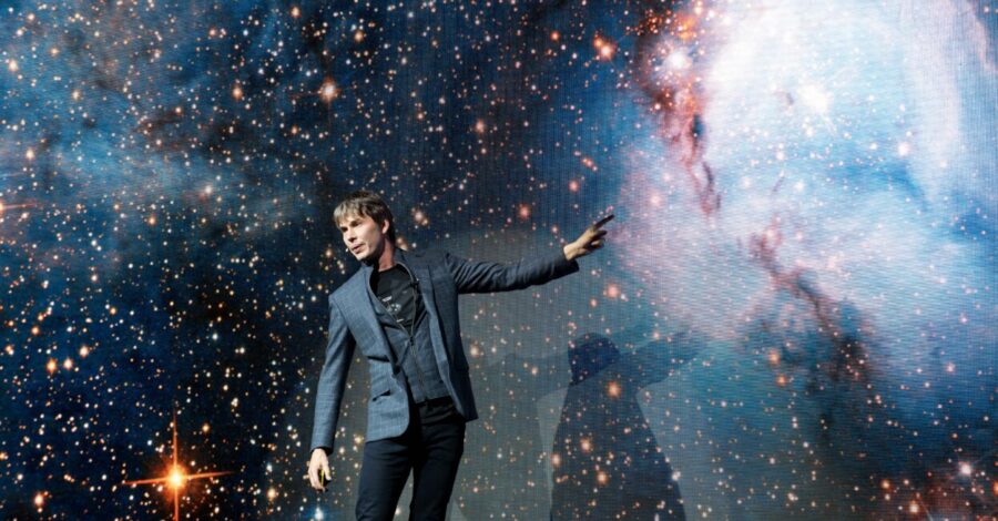 Professor Brian Cox stands on a stage with his left arm pointing to an image of space.
