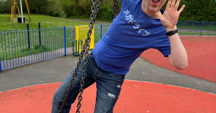 Performer Dommy B stands on a tyre swing in a park wearing a blue t-shirt and jeans. he raises one hand to his mouth as if shouting to be heard.