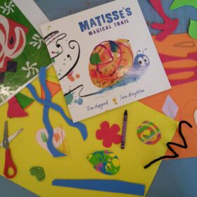 A selection of brightly coloured craft materials including paper,scissors and crayons, and a piece of glossy card with an illustration of a snail printed with the words "Matisse's Magical Trail".