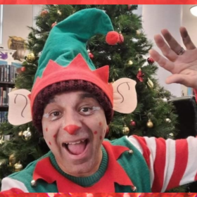 An adult dressed up as an elf with an elf hat and stripey sleeves waving.