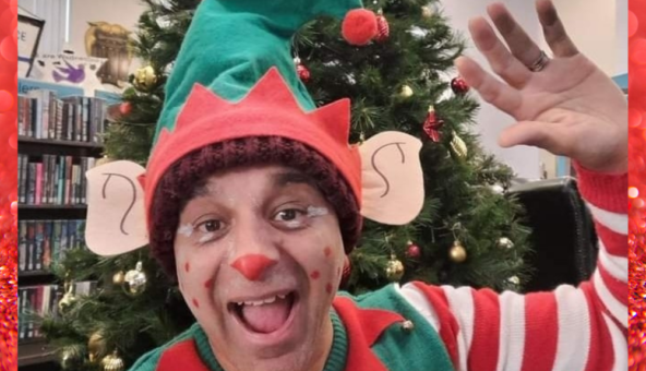 An adult dressed up as an elf with an elf hat and stripey sleeves waving.