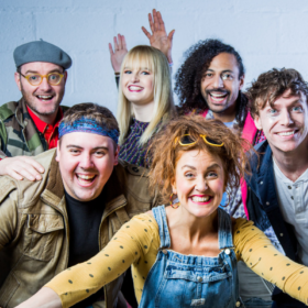 Six member of a pop band stand huddled together smiling.