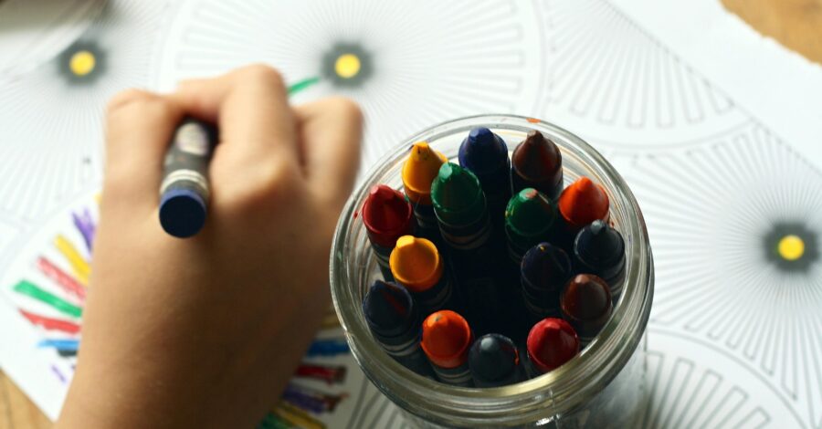 A jar of full of colourful crayons and a child's hand holding a crayon and drawing.