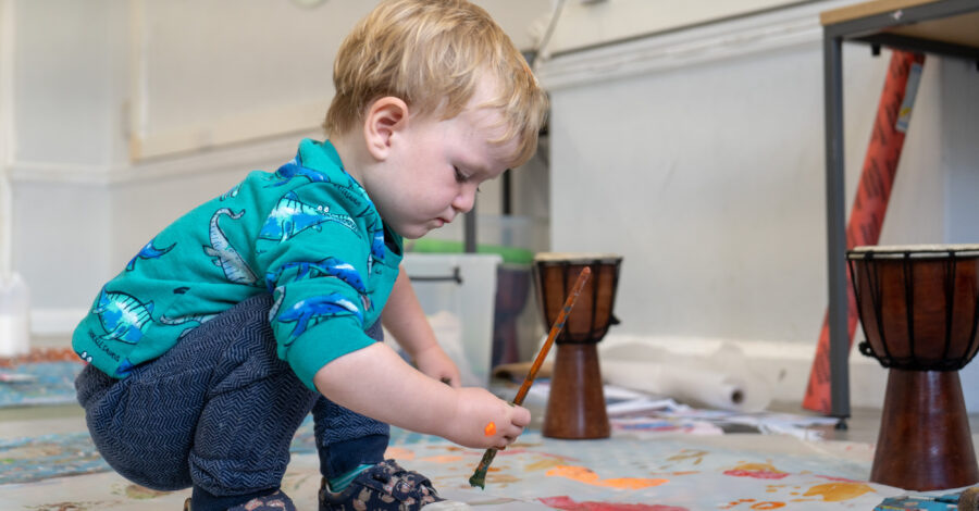 A toddler squats down on the floor holding a paint brush ready to begin painting.