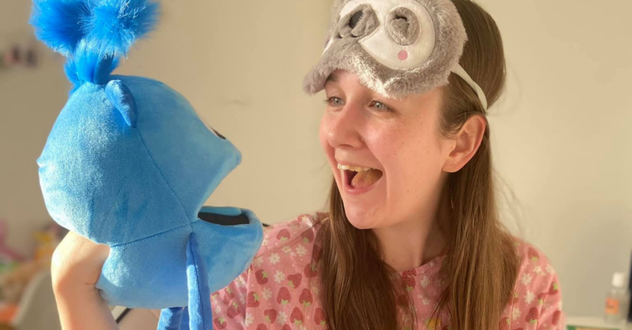 A woman wearing a face mask on the top of her head holds and interacts with a blue plush hand puppet.