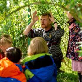 An adult stands in a wooded area with his hands raised at the side of his head as if telling a story. He is surrounded by a group of children.