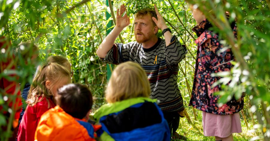 An adult stands in a wooded area with his hands raised at the side of his head as if telling a story. He is surrounded by a group of children.