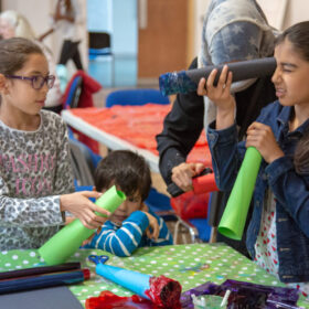 Two young people stand around a crafting table holding green paper tubes.