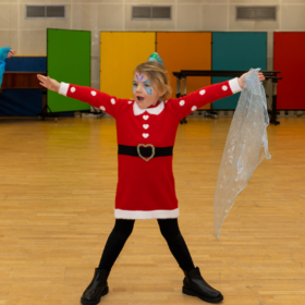 A little girl wearing a santa costume stands with her legs apart and her arms raised in the air.