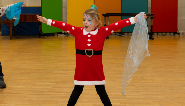 A little girl wearing a santa costume stands with her legs apart and her arms raised in the air.