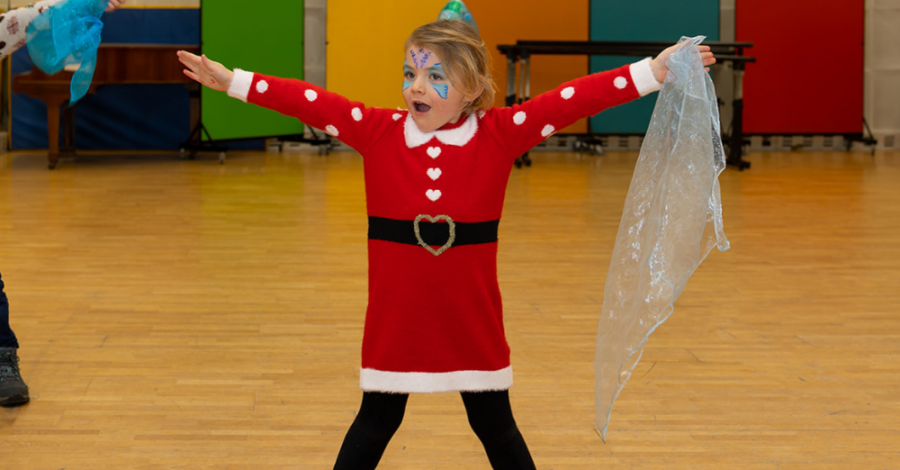 A little girl wearing a santa costume stands with her legs apart and her arms raised in the air.