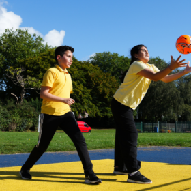 Two young people dressed in yellow t-shirts and black trousers play with an orange ball.