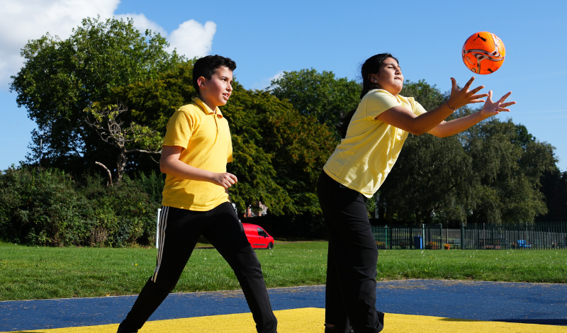 Two young people dressed in yellow t-shirts and black trousers play with an orange ball.