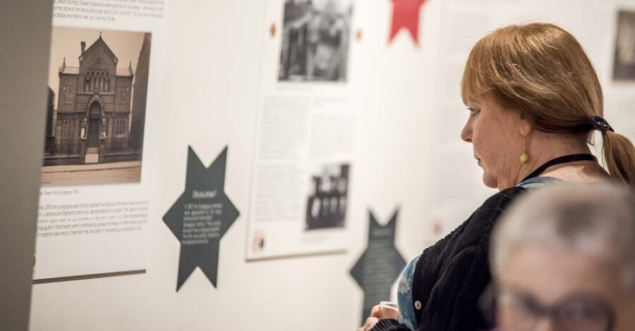 A middle aged woman with light brown hair and a ponytail is looking at a panel exhibition on a white wall. The exhibition consists of big white panels with text and smaller, red and green stars with additional bits of text on them.